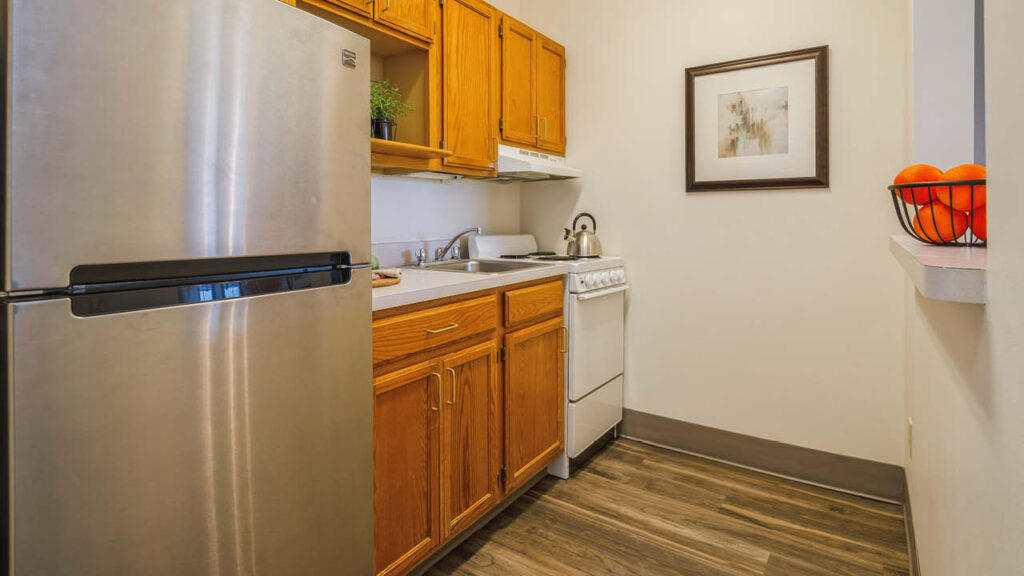 A small kitchen with wood cabinets, a stainless steel refrigerator, a white stove, a kettle, a sink, and a countertop. A framed picture hangs on the wall and a bowl of oranges sits on a ledge.