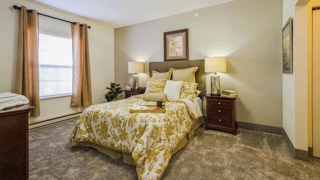 A neatly made bed with yellow floral bedding is centered in a cozy, carpeted bedroom. Two bedside tables with lamps flank the bed. A window with tan curtains and a potted plant are on the left side of the room.