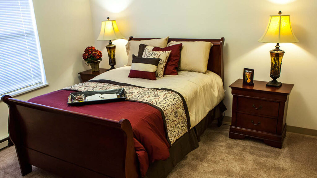 A neatly made bed with cream and burgundy bedding sits between two wooden nightstands, each with a lamp. A tray is on the bed, and a window with closed blinds is on the left. The room has beige carpet and light walls.