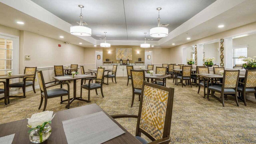 A spacious, well-lit dining room with multiple square tables and patterned chairs, neutral walls, hanging lights, and decorative artwork on the far wall. Each table has a placemat and a small vase with a white flower.