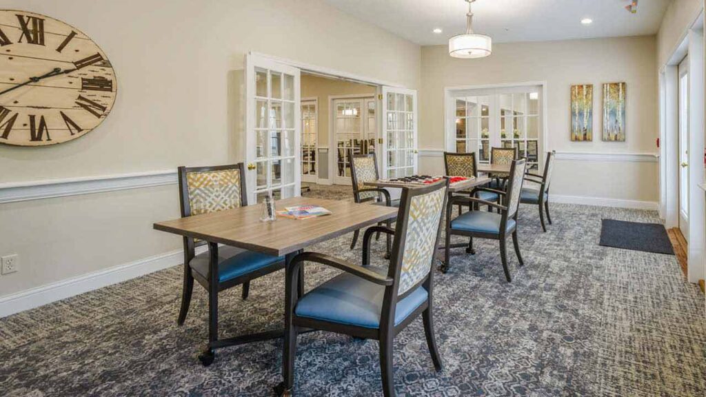 A spacious, well-lit room with tables and chairs set up for games or reading. The room features a large wall clock, patterned carpet, double glass doors, and artwork on the walls.