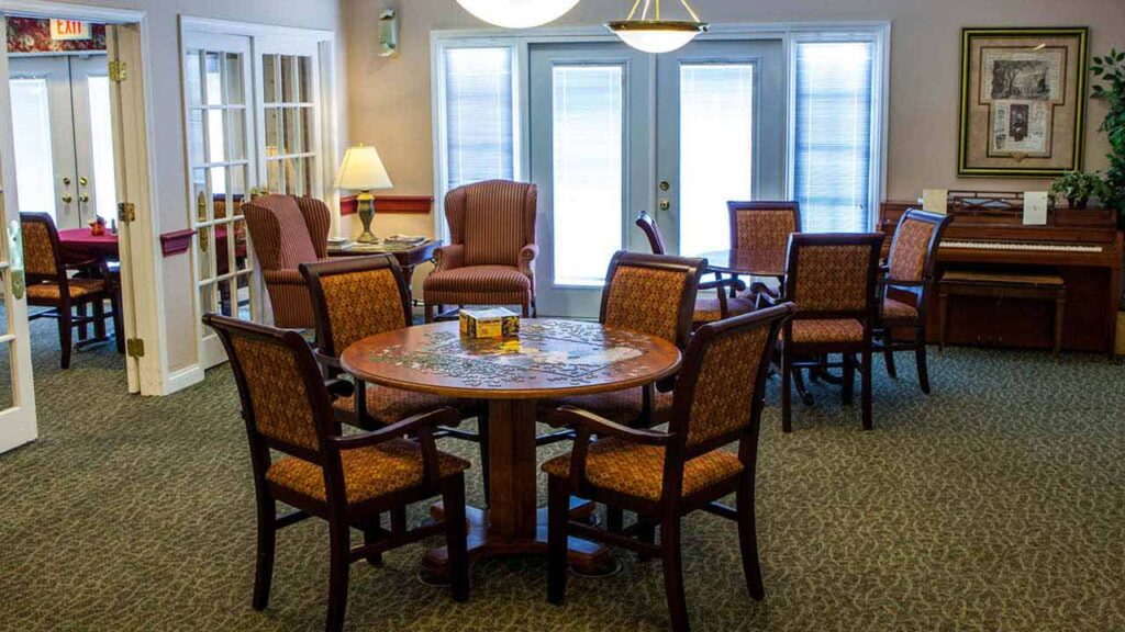A cozy room with round tables and cushioned chairs, two armchairs by a side table with a lamp, a piano against the wall, framed art, and large windows letting in natural light.