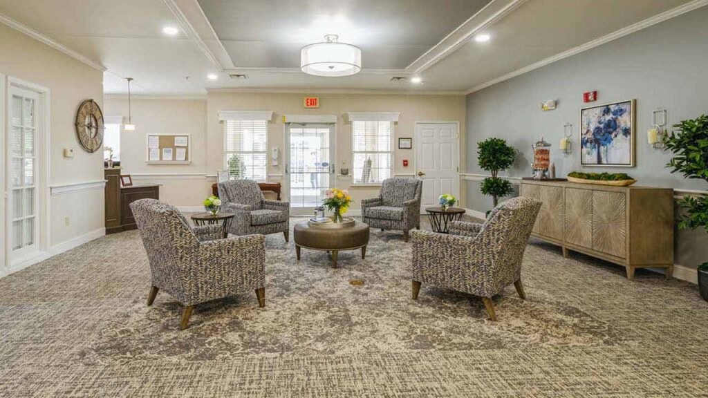 A spacious, well-lit lounge area with four patterned armchairs around a circular ottoman, decorative plants, artwork on the walls, and large windows letting in natural light.