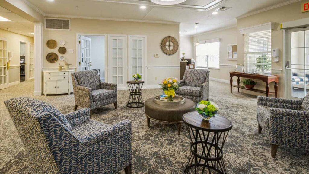 A cozy lounge area with four patterned armchairs, two round side tables, and a round ottoman with flowers, set on a patterned carpet. Natural light fills the room, and decorative items adorn the walls and tables.