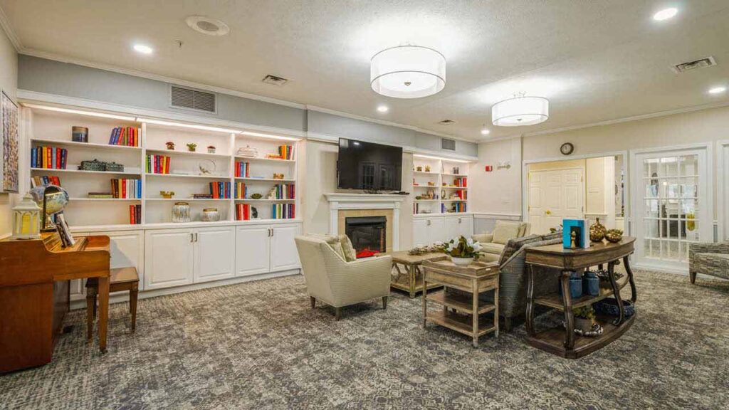A cozy, well-lit living room features armchairs, a coffee table, a piano, and built-in white bookshelves filled with colorful books. A TV is mounted above a fireplace, and the room has modern ceiling lights.