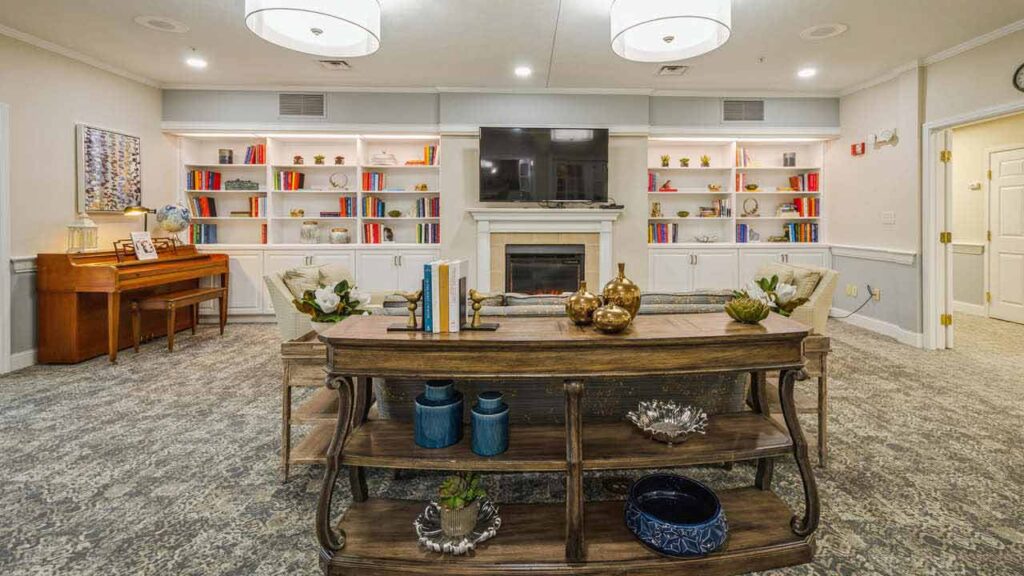 Bright living room with built-in bookshelves, a fireplace, TV, and decorative items. A wooden console table with vases and books stands behind a sofa, and a piano sits against the left wall. The space looks cozy and inviting.