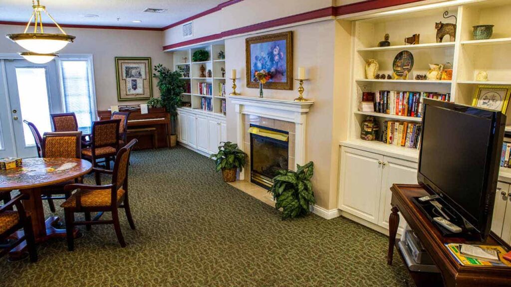 A cozy living room with a dining table and chairs, bookshelves filled with books and decor, a fireplace with plants, a TV, a piano, artwork above the mantel, and large windows letting in natural light.