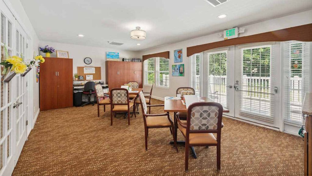 A bright, spacious room with brown carpet, several tables and cushioned chairs, wooden cabinets, a desk with a computer, and large windows with blinds letting in natural light. Paintings and flowers decorate the space.
