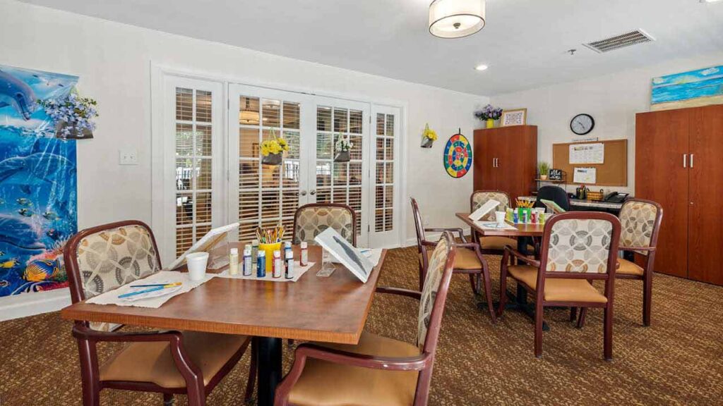 A bright activity room with tables set up for painting, surrounded by chairs. Art supplies and easels are on the tables. Cabinets, artwork, and plants decorate the space. French doors let in natural light.
