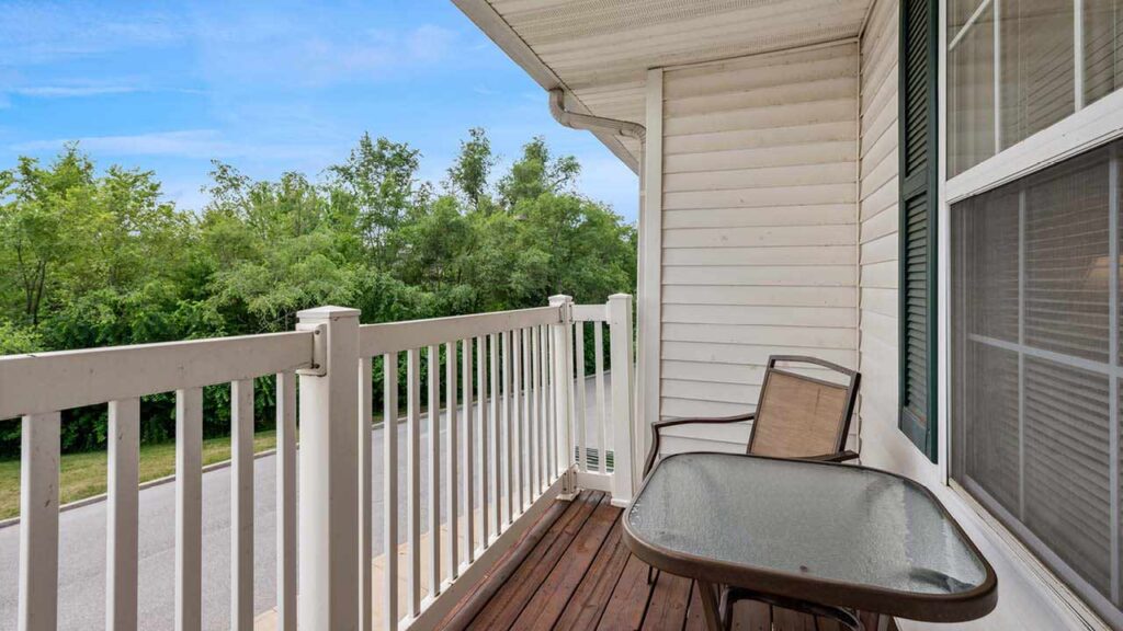 A small balcony with a glass-top table and a single chair, bordered by a white railing, overlooks a quiet road and green trees under a blue sky.