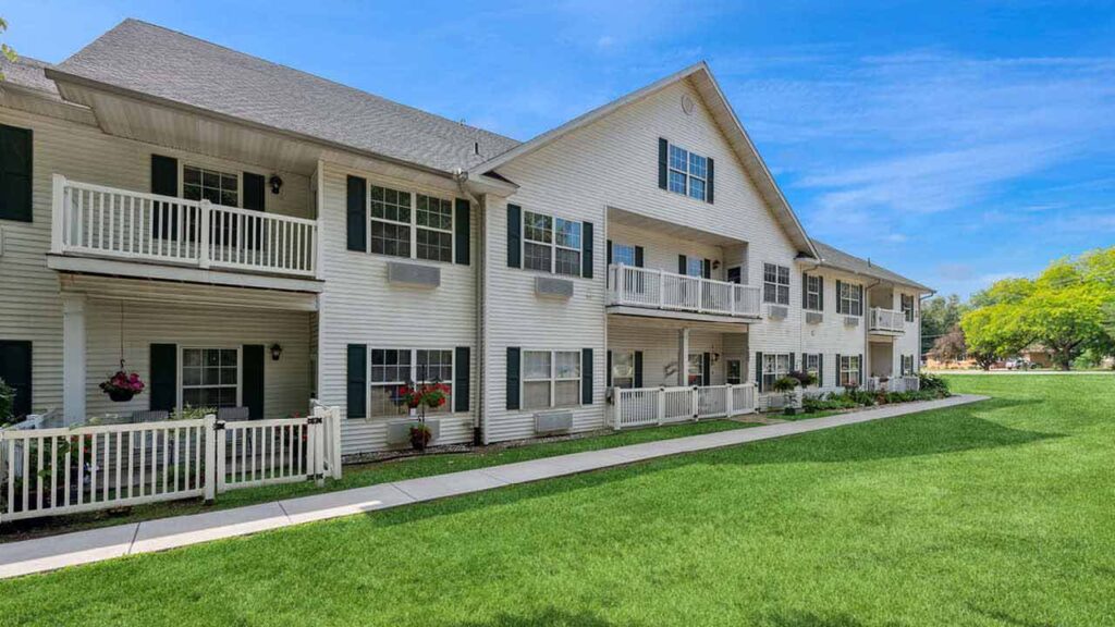 A two-story apartment building with white siding, balconies, and black shutters, surrounded by green grass and a few potted plants, under a bright blue sky.