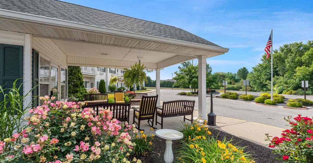 A covered porch with wooden chairs and tables overlooks a parking lot and garden with blooming flowers and greenery. An American flag stands in the background near trees and a clear blue sky.
