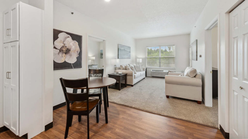 A bright, modern living space with a small dining table and chairs, beige sofas, large windows letting in natural light, and neutral decor including a large flower painting on the wall.