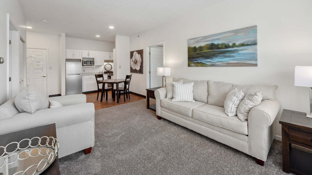 A modern living room with a beige sofa, armchair, and side tables, leading to a dining area with a table and chairs and an open kitchen. Neutral tones and landscape artwork create a cozy, inviting atmosphere.