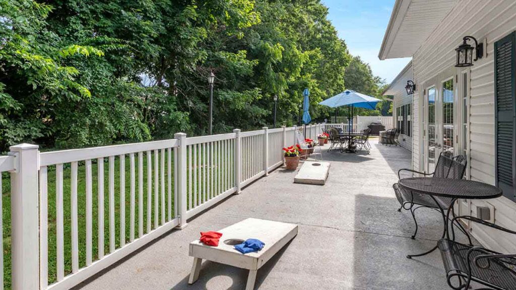 A spacious backyard patio with a white fence, outdoor tables, chairs, flower pots, an umbrella, and cornhole boards. Lush green trees border the yard, and the patio is attached to a white house.