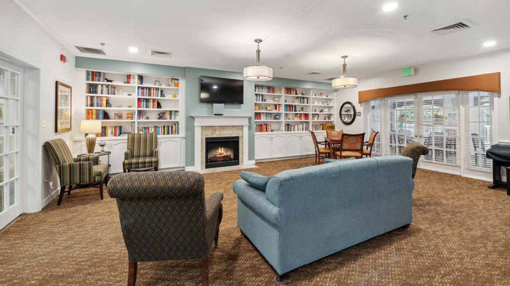 A cozy lounge with armchairs, a blue sofa, a fireplace, bookshelves, a wall-mounted TV, round tables, and large windows letting in natural light, creating a welcoming atmosphere.