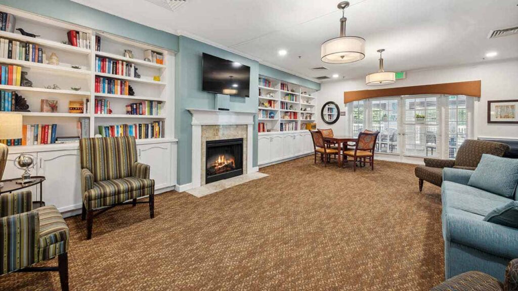 A cozy lounge with a fireplace, bookshelves, armchairs, a sofa, and a table with chairs near large windows letting in natural light.