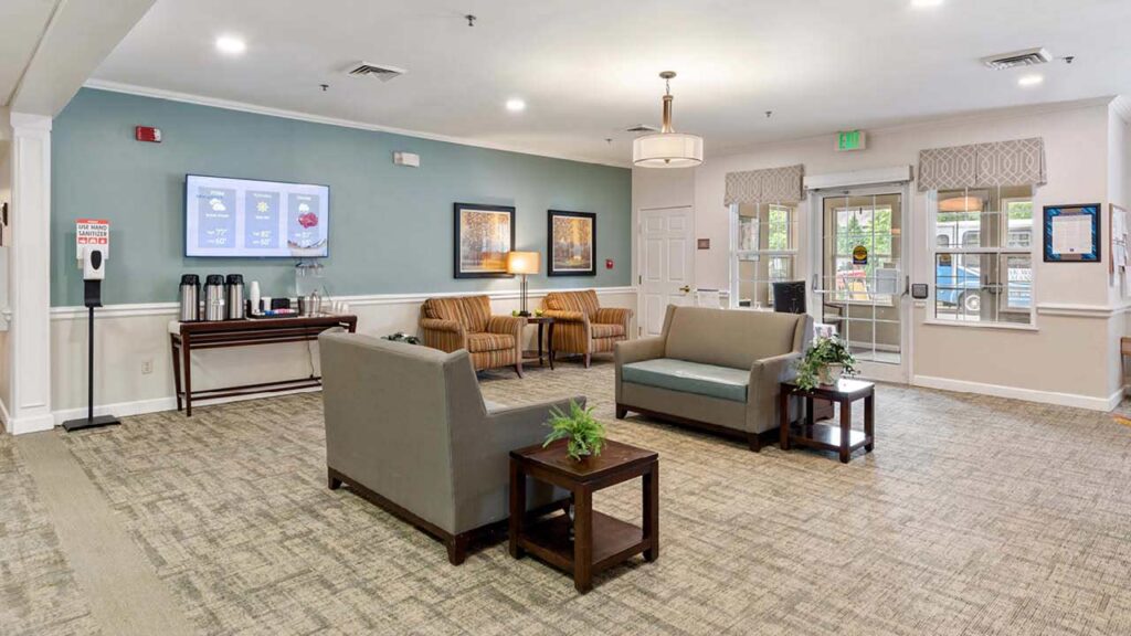 A bright, spacious lobby with two gray sofas, two striped armchairs, coffee tables with plants, a refreshment station, wall art, and large windows near the entrance letting in natural light.
