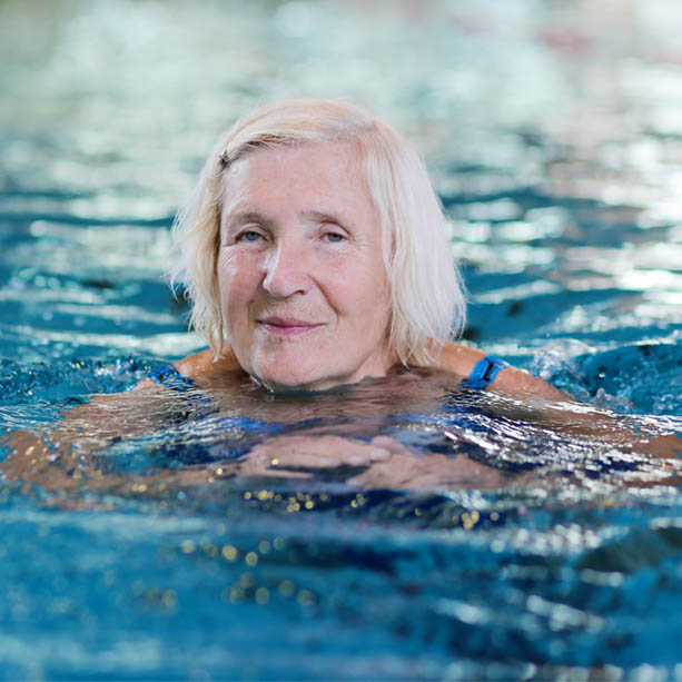 An elderly woman with short white hair is swimming in a pool, her shoulders submerged and wearing a blue swimsuit, with a gentle smile on her face and water reflecting around her.
