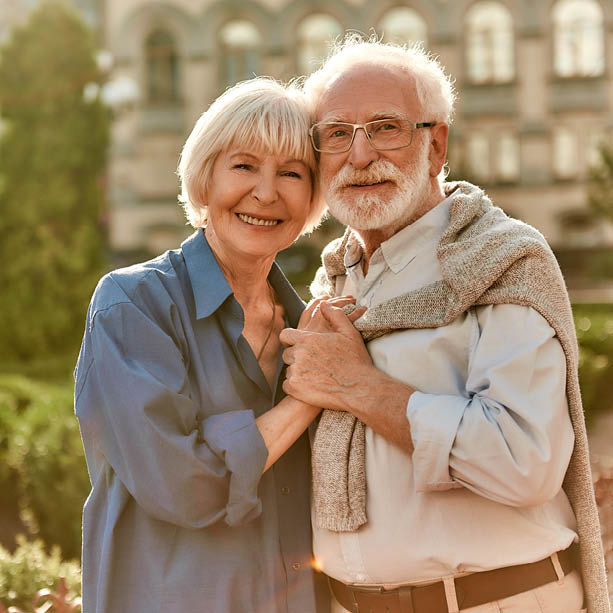 An elderly couple stands outdoors, smiling and holding hands. The man has white hair, a beard, glasses, and a sweater draped over his shoulders. The woman has short, light hair and wears a blue shirt. They appear happy and affectionate.