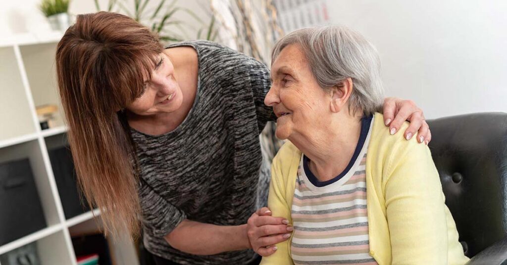 A younger woman smiles and gently embraces an older woman sitting in a chair, both looking at each other warmly in a bright, home-like setting.