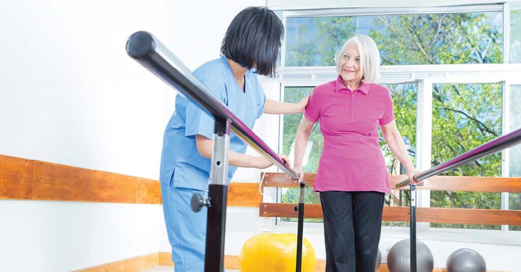 An older woman in a pink shirt walks between parallel bars, assisted by a healthcare worker in blue scrubs, in a bright rehabilitation room with large windows and exercise equipment.