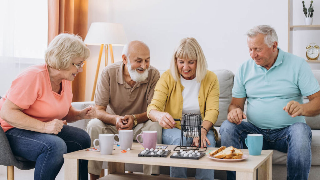 Four older adults sit on a couch and chairs around a coffee table, playing a board game and smiling. The table has mugs and plates with snacks in a cozy, well-lit living room.
