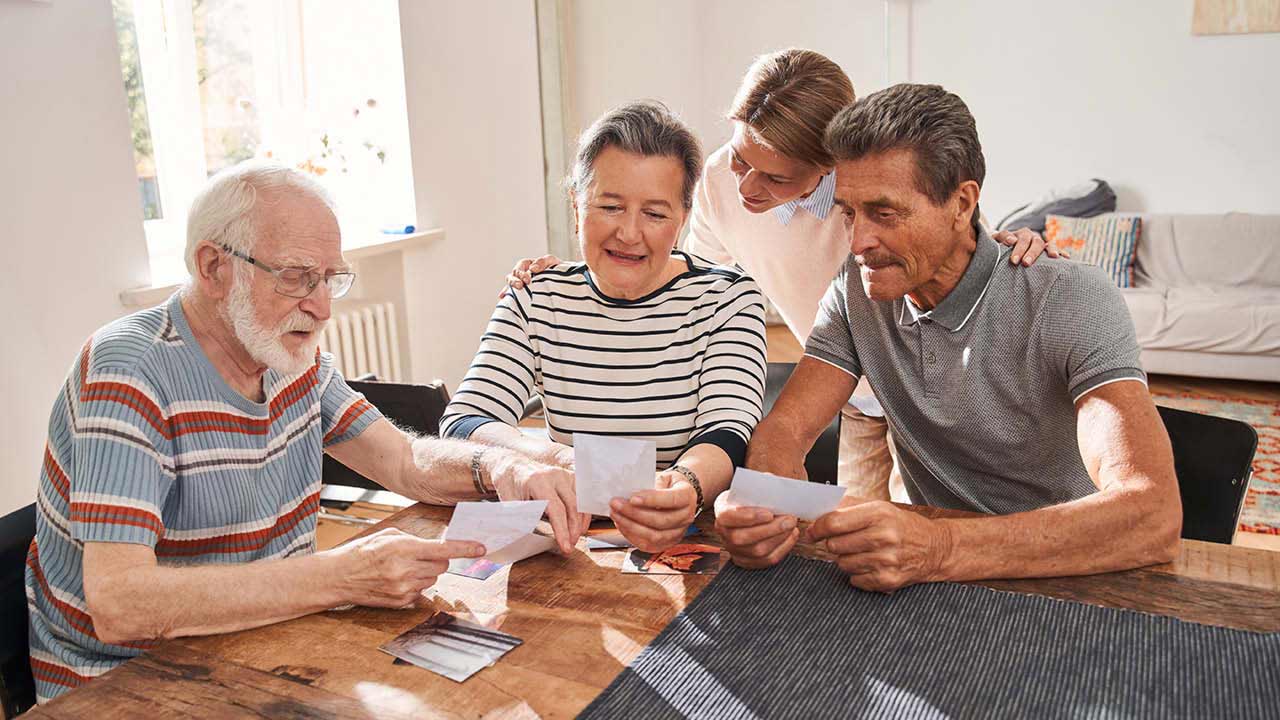 Four older adults sit together at a wooden table, smiling and looking at photographs. A younger person stands behind them, leaning in and joining the moment. The atmosphere appears warm and friendly.