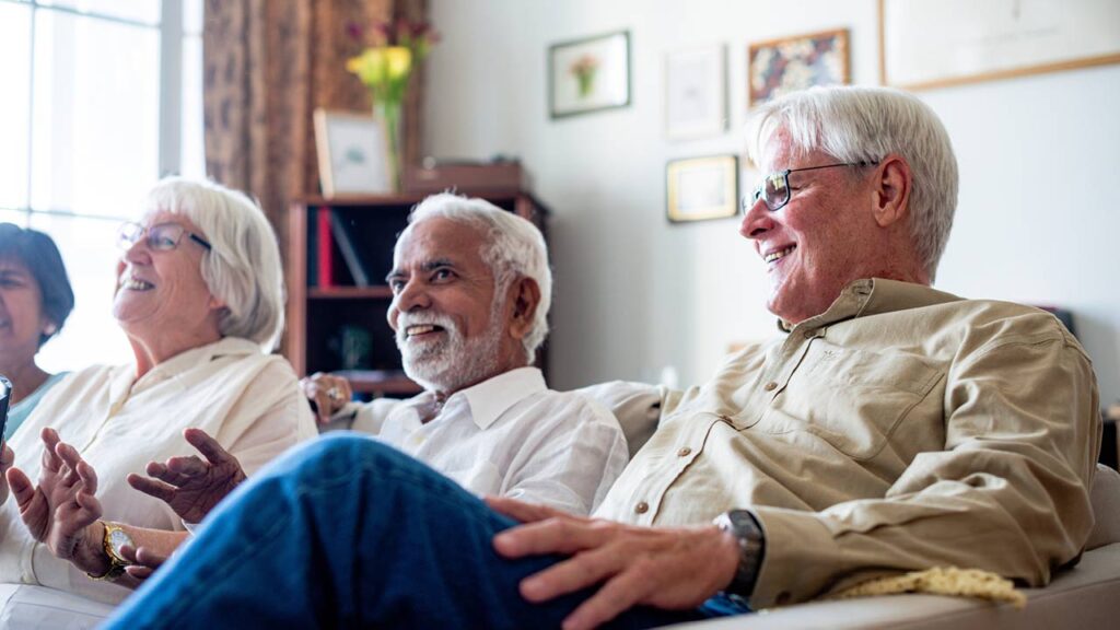 Three older adults sit together on a couch, smiling and laughing in a bright, cozy living room with framed photos and flowers in the background.
