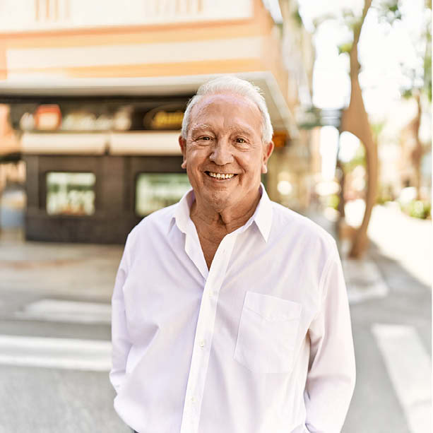 An older man with short white hair smiles warmly while standing outdoors on a sunny street, wearing a white button-up shirt. The background features buildings, trees, and soft, natural light.