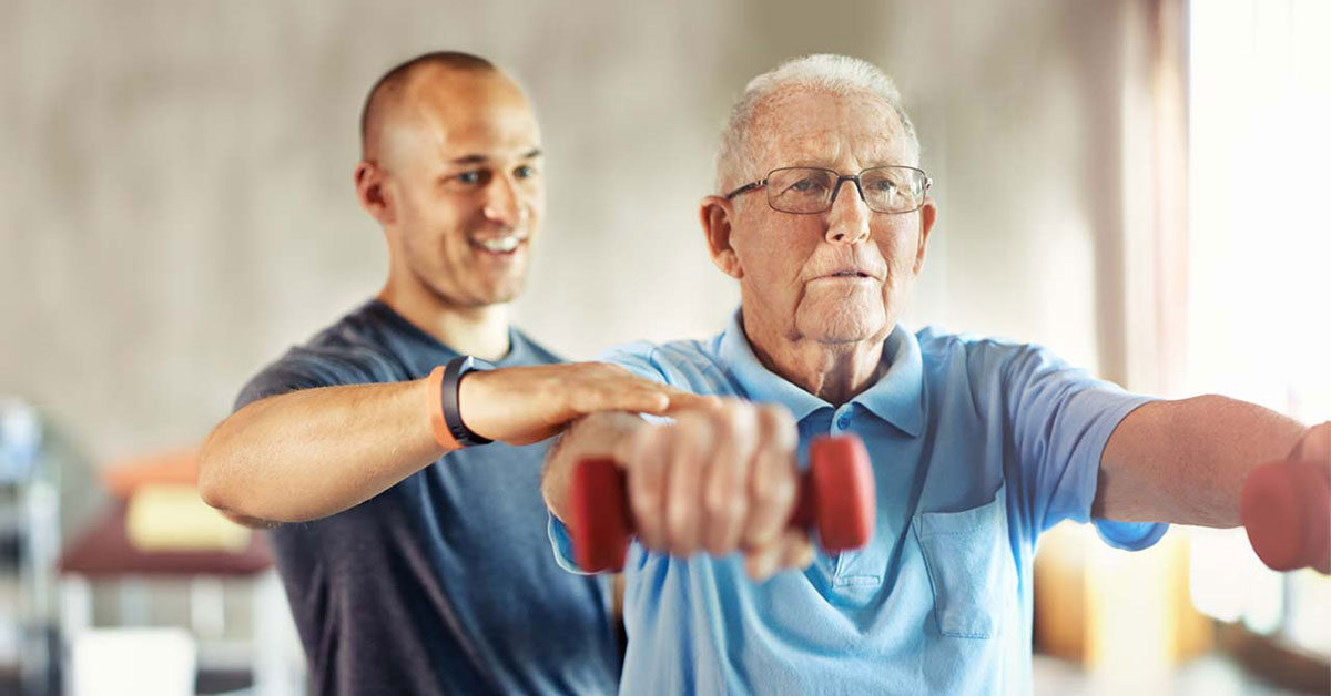 A younger man helps an older man in glasses lift red dumbbells during a workout, guiding his form. Both are indoors with soft natural light in the background.