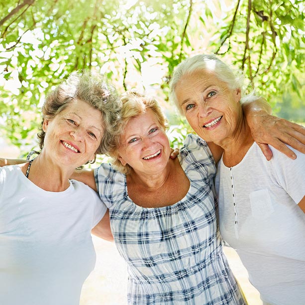 Three older women smiling and embracing each other outdoors under a leafy tree, enjoying a sunny day together.
