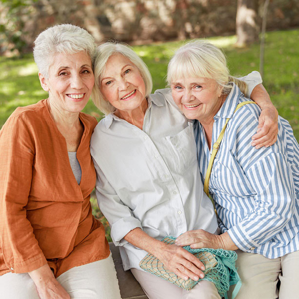 Three older women are sitting closely together on a bench outdoors, smiling and posing for the camera. They appear happy and relaxed, with greenery and trees visible in the background.