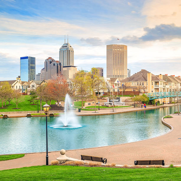 A cityscape view of downtown Indianapolis with modern skyscrapers, a canal with a water fountain, green lawns, trees, park benches, and walking paths under a partly cloudy sky.
