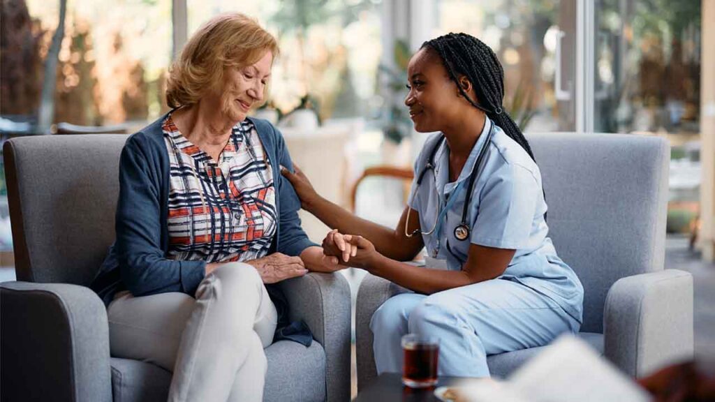 A nurse in scrubs smiles and holds the hand of an older woman sitting in a chair, offering comfort and support in a bright, cozy room.