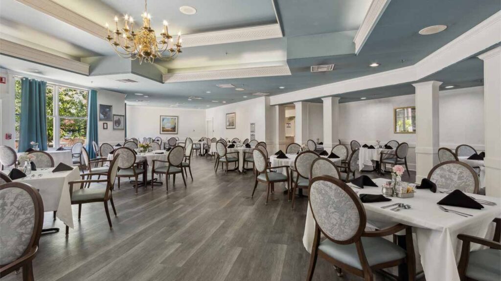 Elegant restaurant dining room with round tables covered in white tablecloths, cushioned chairs, black napkins, chandeliers, and large windows letting in natural light; art decorates the light blue and white interior.