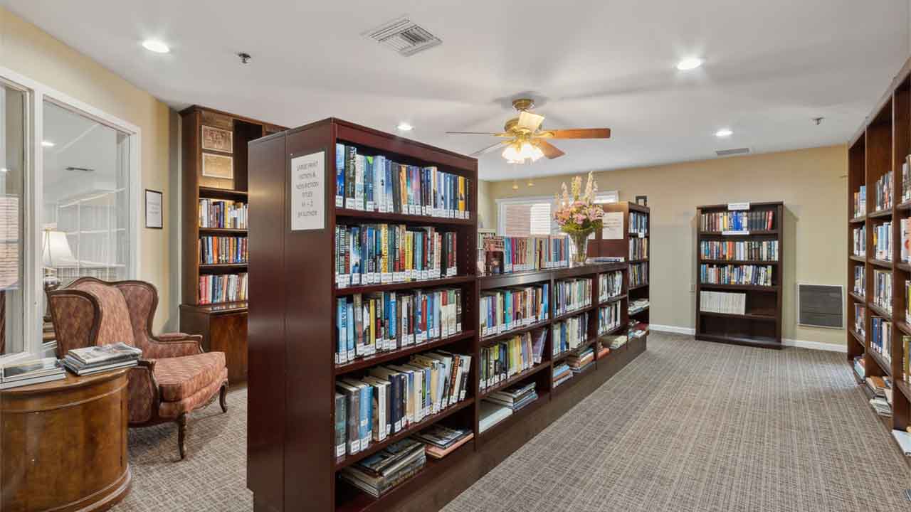 A well-lit library with several bookshelves filled with books, a wooden side table with two armchairs, and a ceiling fan. A window lets in natural light, and flowers decorate the top of a bookshelf.