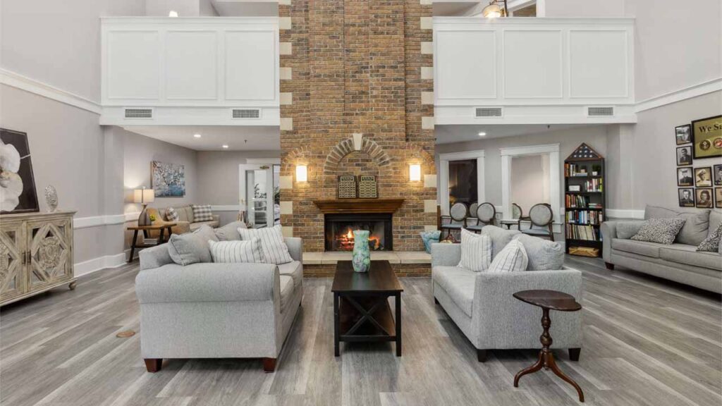 A spacious living room with light gray sofas, a dark coffee table, and a large brick fireplace as the focal point. The room features wood flooring, soft lighting, and decorative accents throughout.