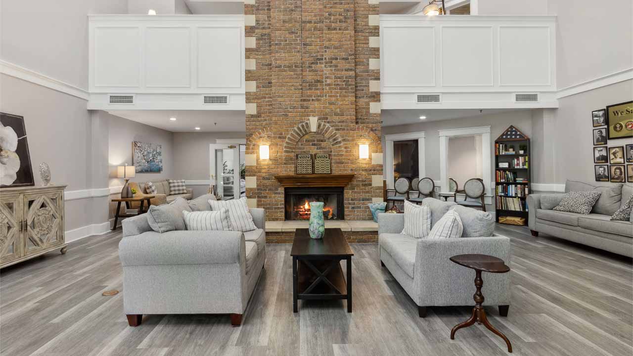 A spacious living room with light gray sofas, a dark coffee table, and a large brick fireplace as the focal point. The room features wood flooring, soft lighting, and decorative accents throughout.