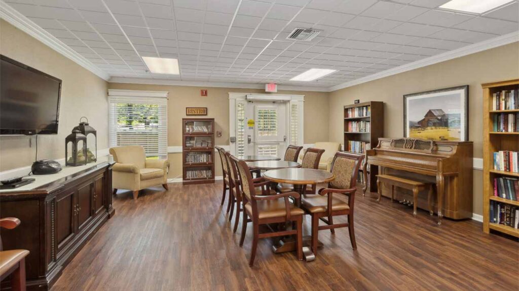 A cozy communal room with wooden floors, a round table with chairs, an armchair, a piano, bookshelves, a TV, and large windows letting in natural light.