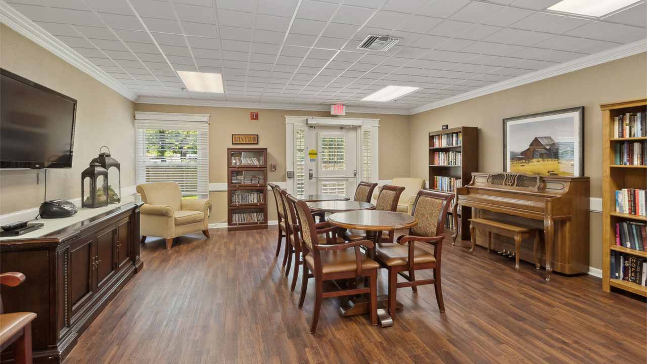 A cozy communal room with wooden floors, a round table with chairs, an armchair, a piano, bookshelves, a TV, and large windows letting in natural light.