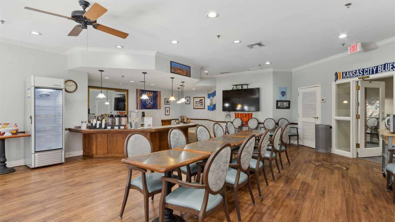 A spacious dining area with wooden floors, several tables and chairs, a bar counter with drinks, and sports memorabilia on the walls, including a "Kansas City Blue" banner. The room is brightly lit with ceiling lights.
