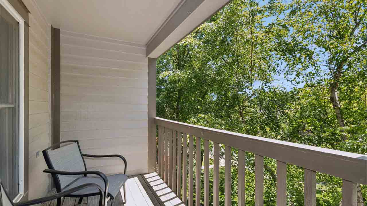 A small balcony with two chairs, wooden railings, and a light-colored wall overlooks leafy green trees under a bright blue sky. Sunlight casts shadows on the floor.