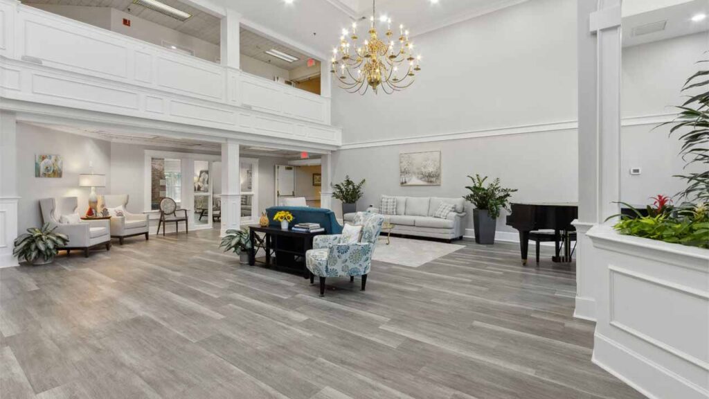 Bright, spacious lobby with light wood flooring, white walls, seating areas with sofas and chairs, indoor plants, a black grand piano, chandelier, and large windows allowing natural light.