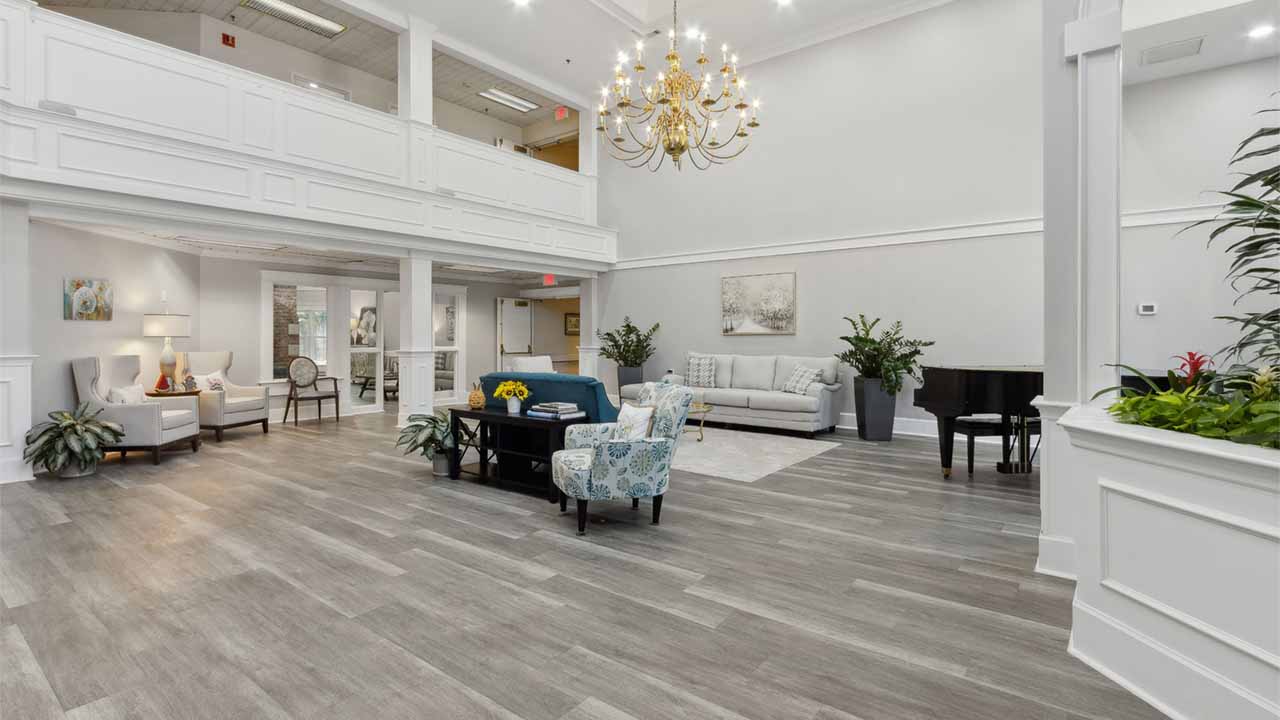 Bright, spacious lobby with light wood flooring, white walls, seating areas with sofas and chairs, indoor plants, a black grand piano, chandelier, and large windows allowing natural light.