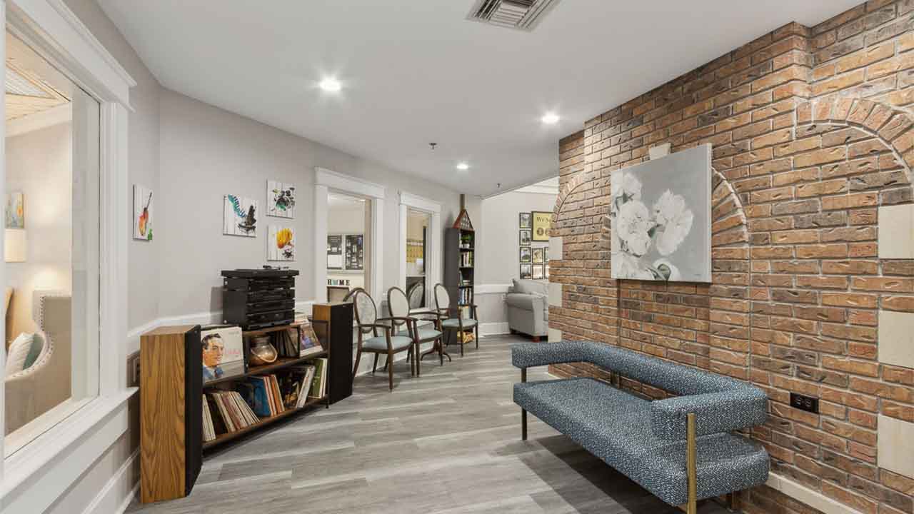 A cozy room with a brick accent wall, blue bench, record player, vinyl records, and art on the walls. Nearby, there's a table with chairs and a window looking into another softly lit room.