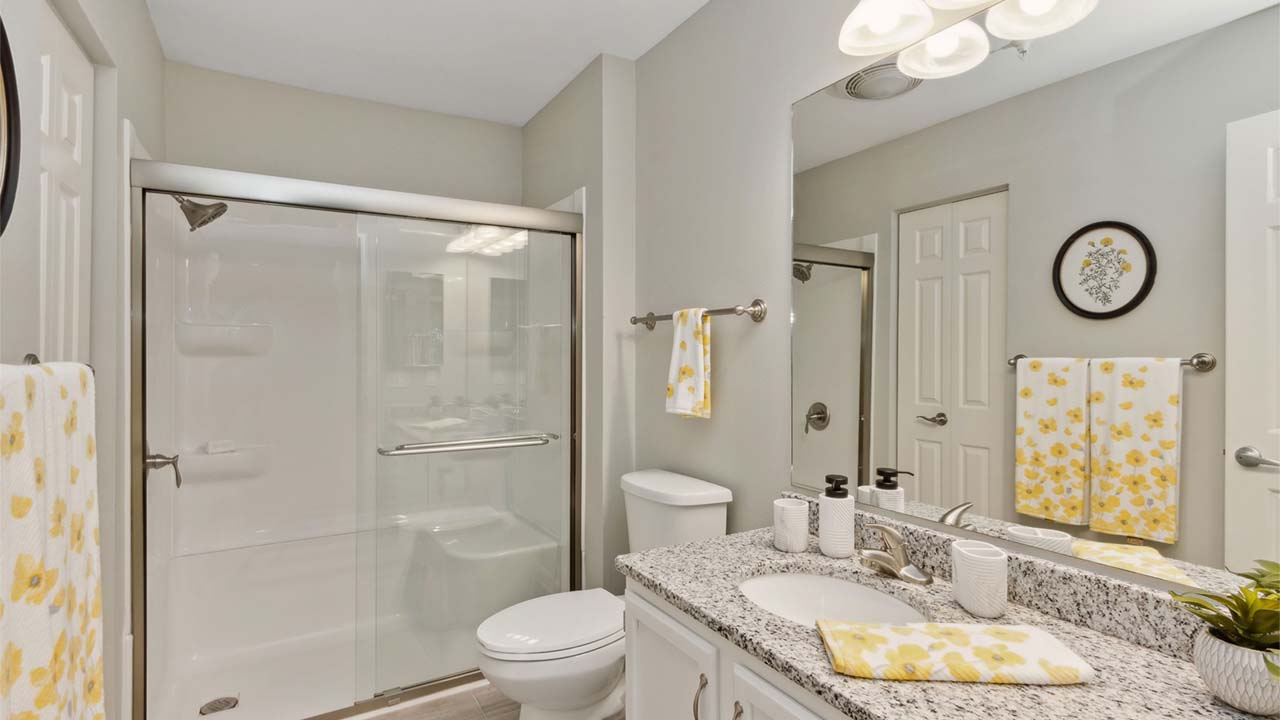 Modern bathroom with a glass-enclosed shower, granite countertop, white cabinetry, and yellow floral towels and decor. A round floral artwork hangs on the wall above the toilet. The overall color scheme is white and gray.