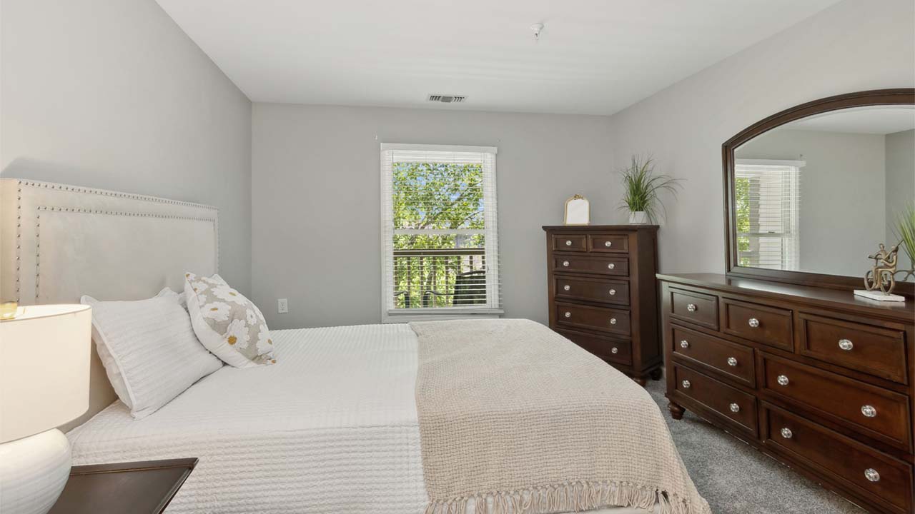A bright bedroom with a large bed, white bedding, and beige throw blanket. There is a wooden dresser with a mirror and a chest of drawers, both decorated with plants and accessories. A window lets in natural light.