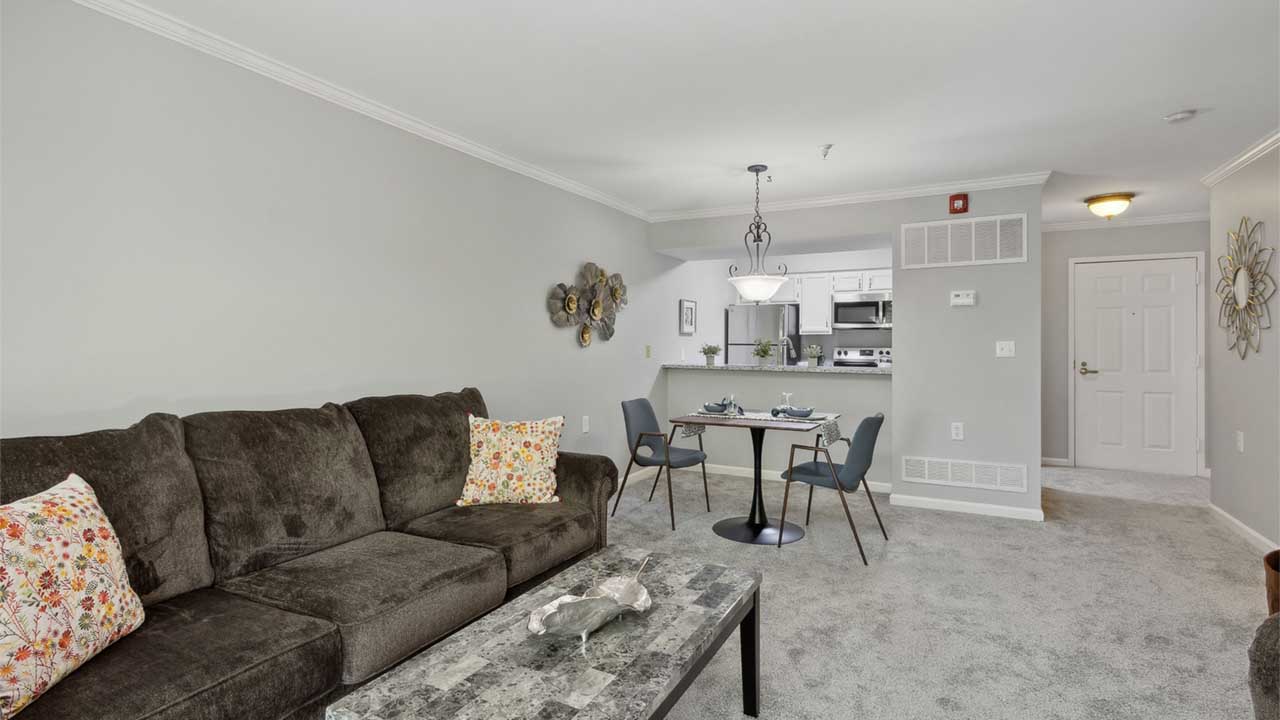A modern living room with a dark sofa, floral pillows, and a marble coffee table. A small dining table with two chairs is near an open kitchen. The space has gray walls and carpet, with decorative wall art.