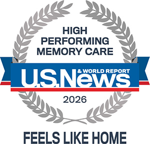 A circular laurel badge with text: "High Performing Memory Care, U.S. News & World Report, 2026." Below the badge reads, "Feels Like Home." A blue ribbon crosses the center behind the U.S. News logo.